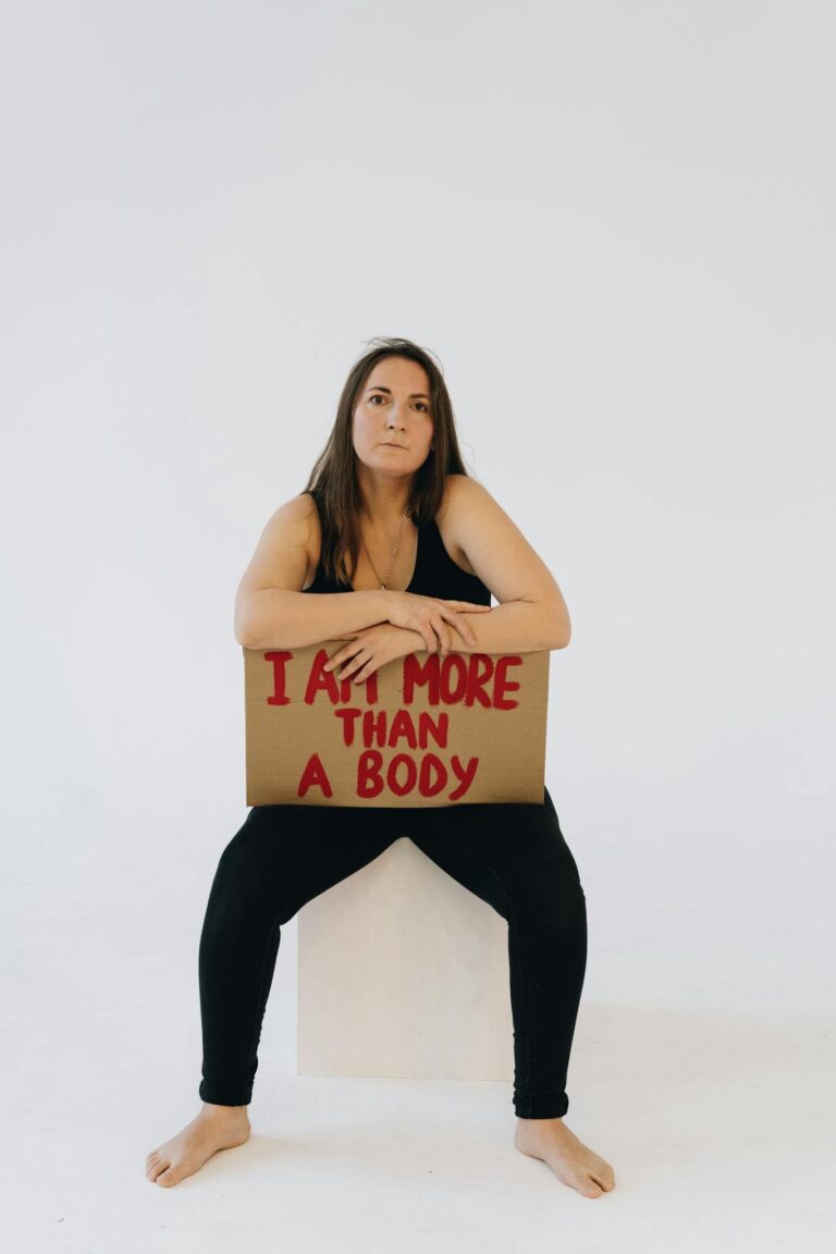 Woman seated with a cardboard sign promoting body positivity and empowerment.